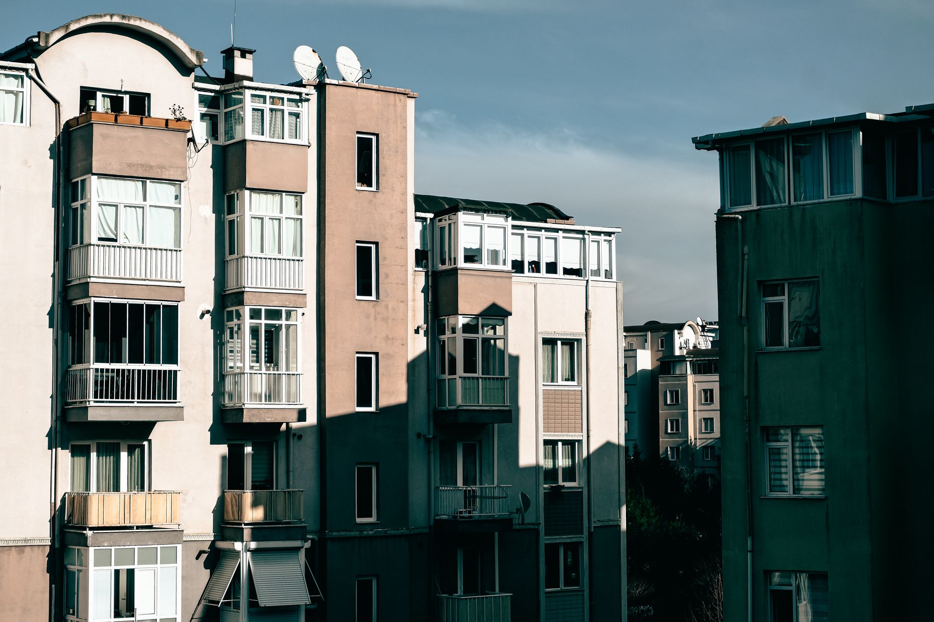 Façades d’immeubles d’habitation avec balcons vitrés, fenêtres et antennes paraboliques sous un ciel bleu, en lumière rasante.