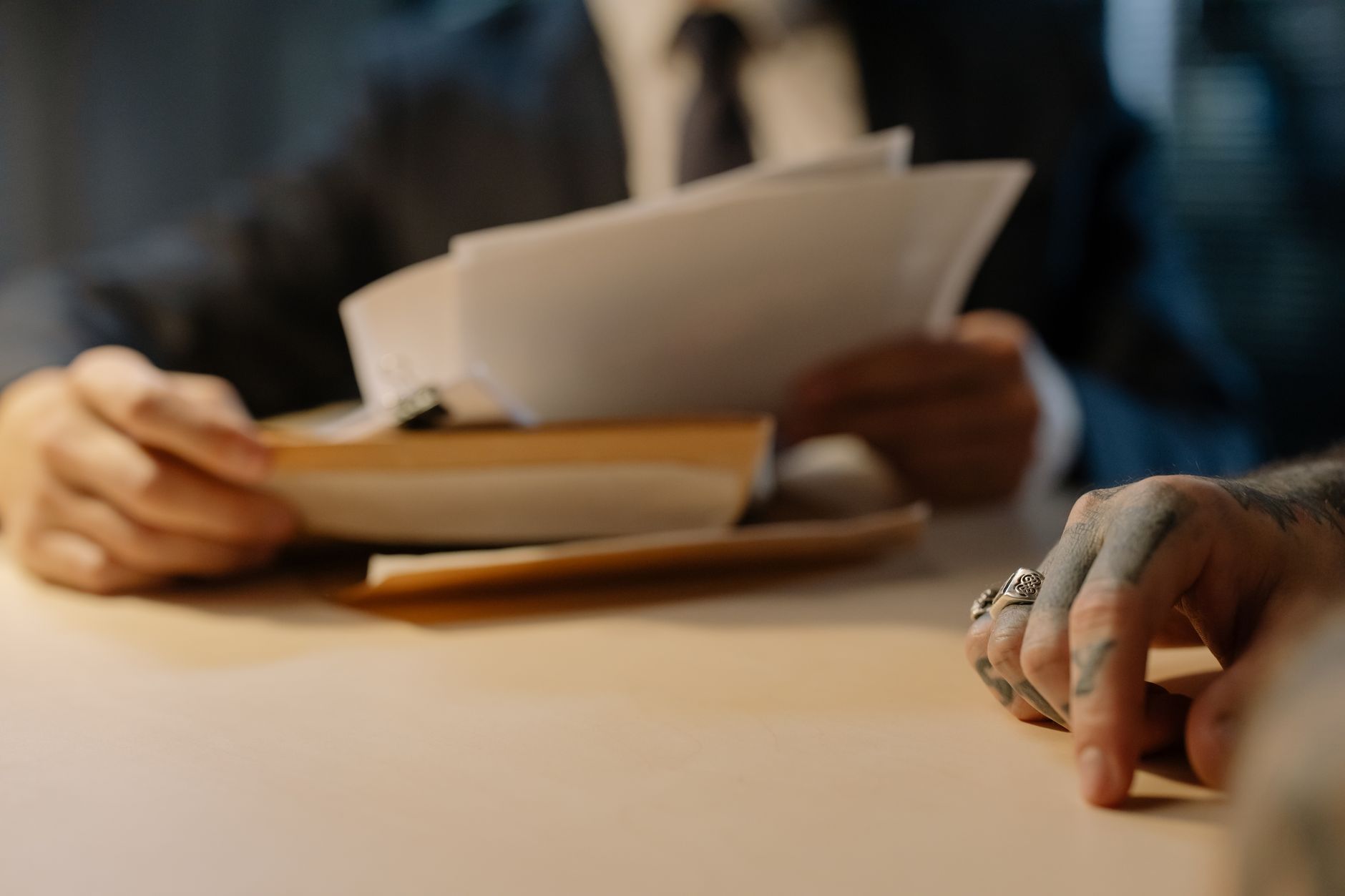 Une personne en costume feuillette des documents sur une table, face à une main tatouée ornée de bagues posée au premier plan.